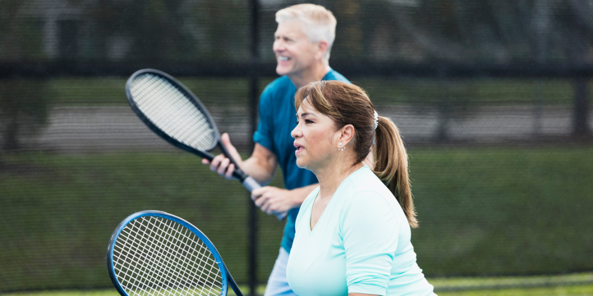 A female player in the foreground and a male player in the background hold tennis racquets.