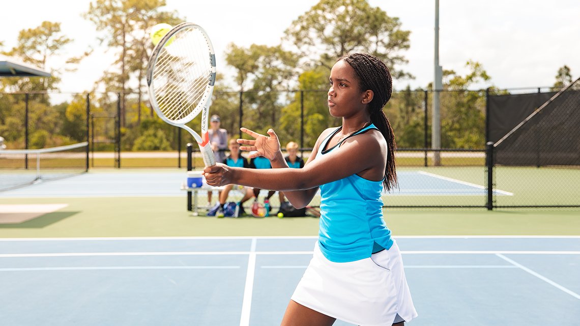 Illustrating the tennis scoring rules for tiebreak games, is a high school tennis player serving during a match, with her team watching in the background.