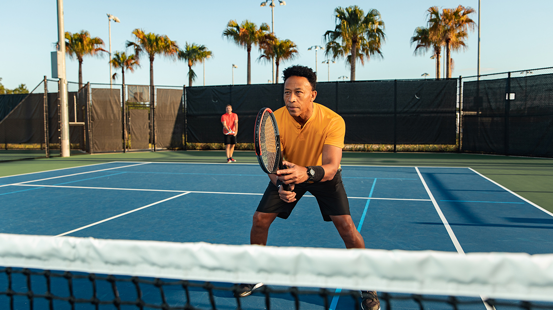 Man holding a tennis racquet near the net, eyeing the ball, illustrating tennis rules for singles and doubles matches on various court surfaces.