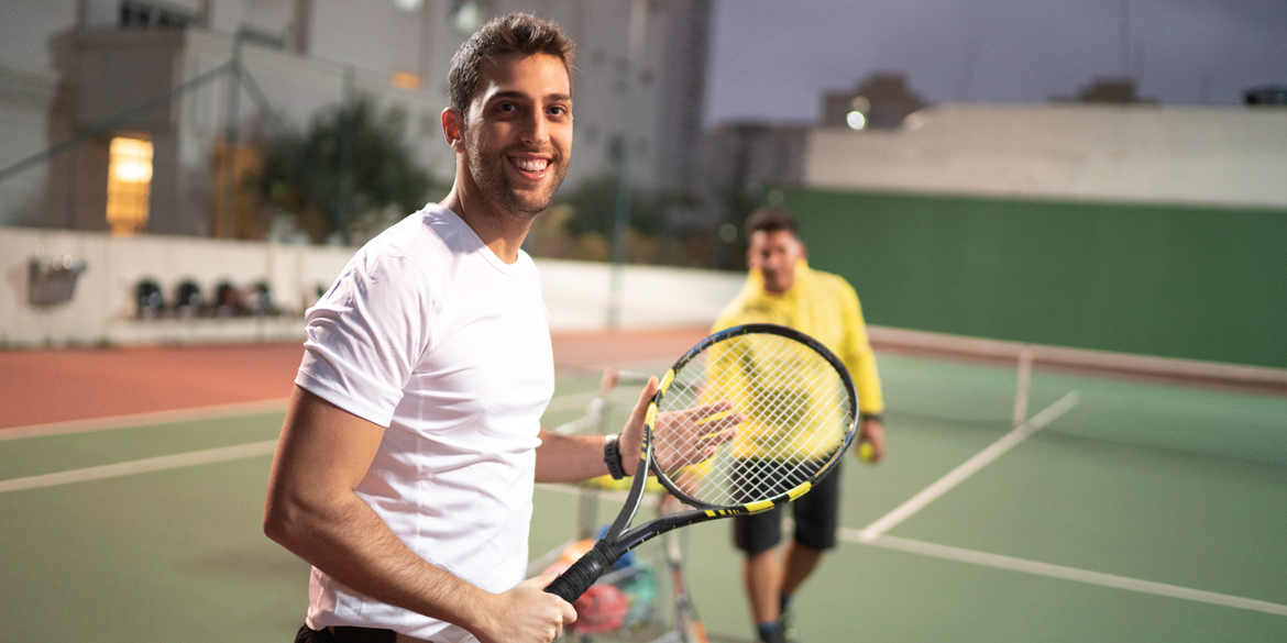 Two men on a tennis court, one smiling and holding a racquet, promoting safe play practices for adult tennis players to foster a positive environment.