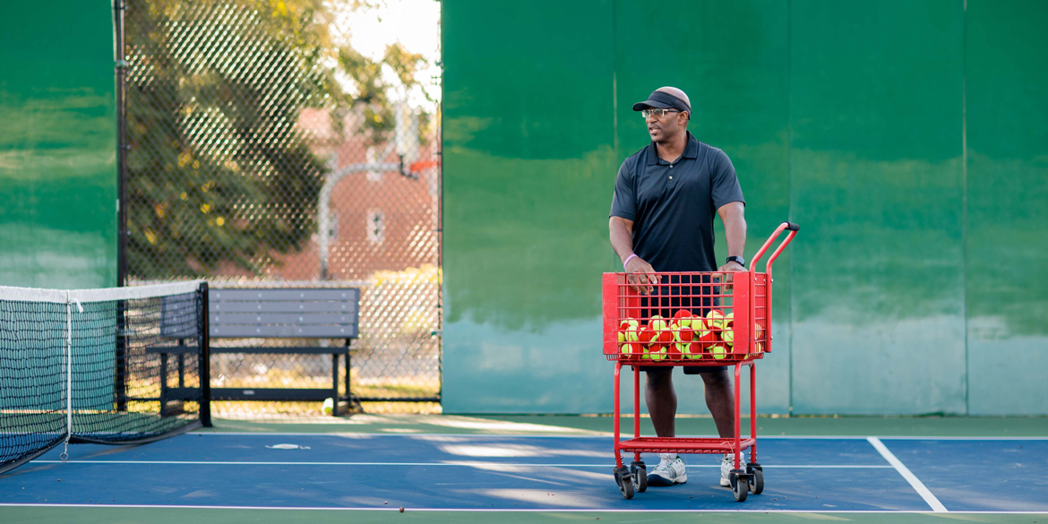 Man on a tennis court with tennis balls in a ball hopper, representing facilities promoting Safe Play to ensure a safe environment for all players.